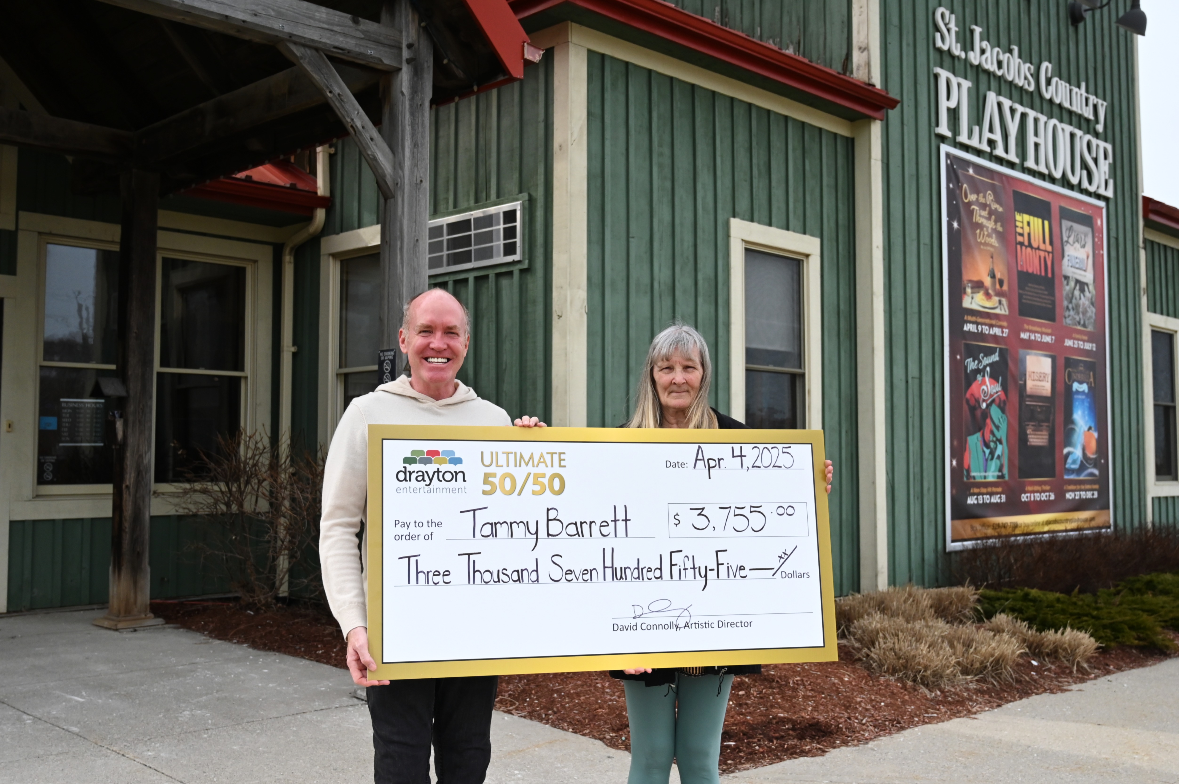 Two smiling individuals stand outside the St. Jacobs Country Playhouse, holding a large cheque made out to Tammy Barrett for $3,755.00 from Drayton Entertainment's 50/50 draw, dated April 4, 2025. The cheque is signed by David Connolly, Artistic Director.